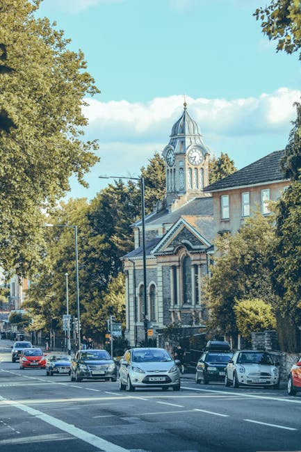 A street scene in Slade Green showing parked cars along the pavement with a mixture of traditional and modern buildings, including a prominent clock tower with a spire and clock face, partly obscured by leafy trees. The road features multiple vehicles, such as small vans and passenger cars, indicating ongoing or upcoming home relocation or furniture transport activities. The pavement area is clear, and the weather appears bright with a partly cloudy sky. This setting reflects a typical urban environment suitable for house removals or moving services, with visible elements such as parking arrangements and surrounding architecture as seen in images related to relocation logistics, demonstrating the environment where Man with Van Slade Green operates.