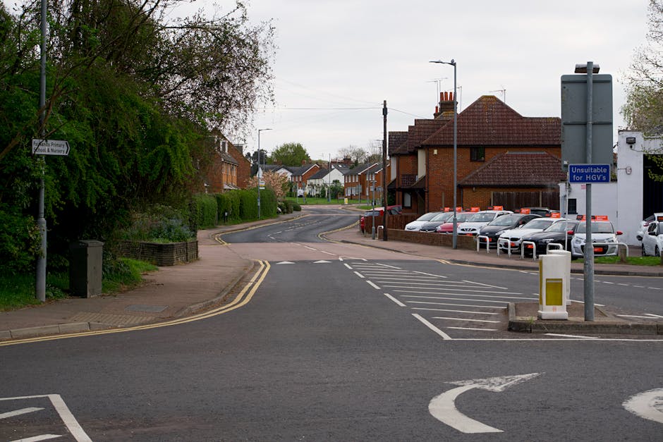 A street scene in Slade Green showing parked cars along the pavement with a mixture of traditional and modern buildings, including a prominent clock tower with a spire and clock face, partly obscured by leafy trees. The road features multiple vehicles, such as small vans and passenger cars, indicating ongoing or upcoming home relocation or furniture transport activities. The pavement area is clear, and the weather appears bright with a partly cloudy sky. This setting reflects a typical urban environment suitable for house removals or moving services, with visible elements such as parking arrangements and surrounding architecture as seen in images related to relocation logistics, demonstrating the environment where Man with Van Slade Green operates.