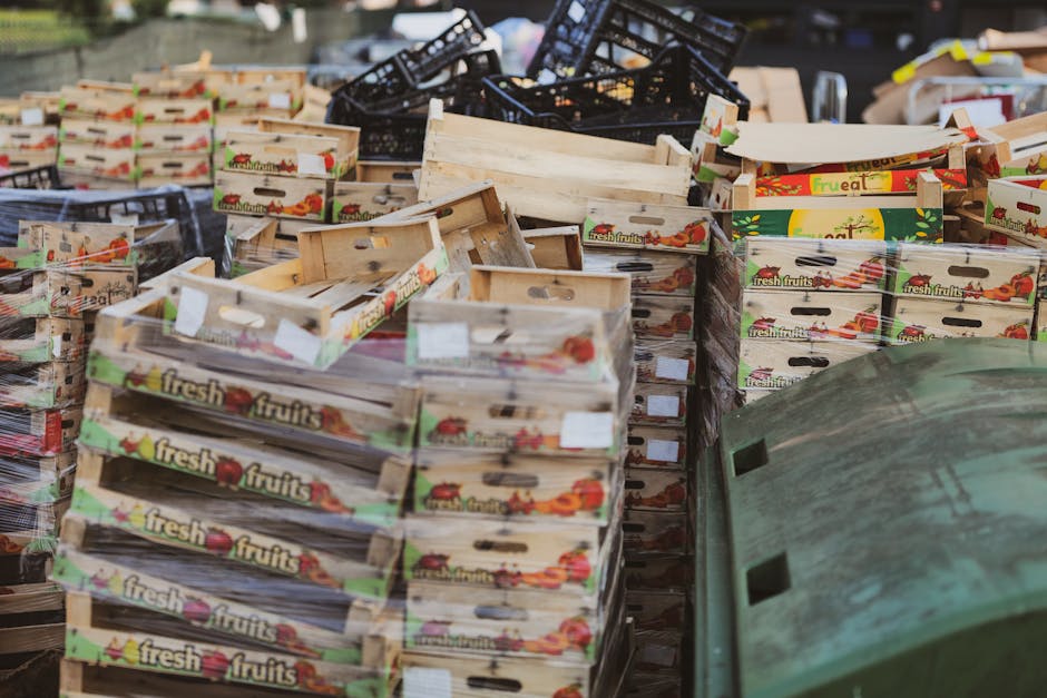 A large number of wooden crates labeled with 'fresh fruits' are stacked in an outdoor area, arranged on a pavement next to a green wheelie bin. The crates are made of light-colored wood with cut-out h