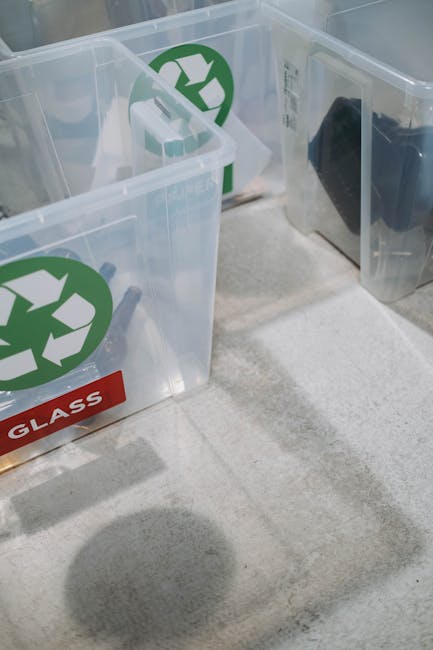 Three transparent plastic recycling bins with green recycling symbols and a red label marked 'GLASS' are aligned side by side on a light grey carpeted floor inside a property. The bins contain various recyclable materials, including cardboard packages and bottles. The rightmost bin has a black plastic object inside, possibly a container or packaging. The environment is well-lit, and the bins are positioned near a doorway, suggesting they are ready for collection or transportation during a home relocation or house clearance process. Occasionally, [COMPANY_NAME] handles such removals, including packing and loading tasks, as part of their house moving services related to house clearance regulations like Bexley Council's bulky waste rules as described on the webpage [PAGE_TITLE].