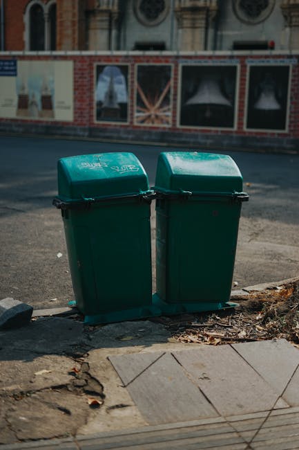 Two green plastic wheelie bins with closed lids are positioned upright on the pavement near the edge of a street, in front of a curb and concrete sidewalk slabs. The bins are placed close to each other, slightly leaning toward one another, with some dirt and debris visible around their bases. Behind the bins is an asphalt road with a faintly visible traffic marking. In the background, there is a construction site or building with red brick walls, large windows, and decorative architectural details, partially obscured by a temporary wooden fence. Natural daylight illuminates the scene, suggesting daytime. This image is relevant to house removals and logistics, reflecting possible waste disposal or clearing process during home relocation or property clearance managed by Man with Van Slade Green, part of their removals service for handling bulky waste, including garden or household rubbish during house clearances in the DA8 area of Bexley Council.