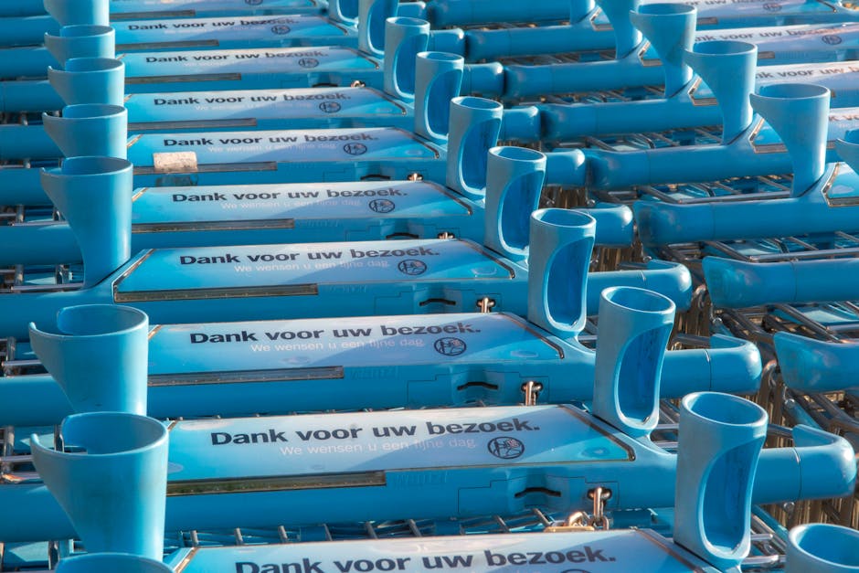 Multiple blue plastic shopping trolleys arranged in rows outside a property, with some positioned on a pavement and others possibly inside a loading area. Each trolley has a printed message in Dutch that reads 'Dank voor uw bezoek,' which translates to 'Thank you for your visit,' indicating they may be for customer use. The trolleys are made of durable plastic with metal handles and wheels, some of which are visible at the bottom. The scene is well-lit, capturing the trolleys' clean and organized appearance, and may be part of a commercial setting such as a supermarket or retail store involved in a house removal or relocation process. Occasionally, the trolleys are associated with packing or moving activities, relevant to [COMPANY_NAME]'s services in removals, loading, and transportation logistics for house or business moves.
