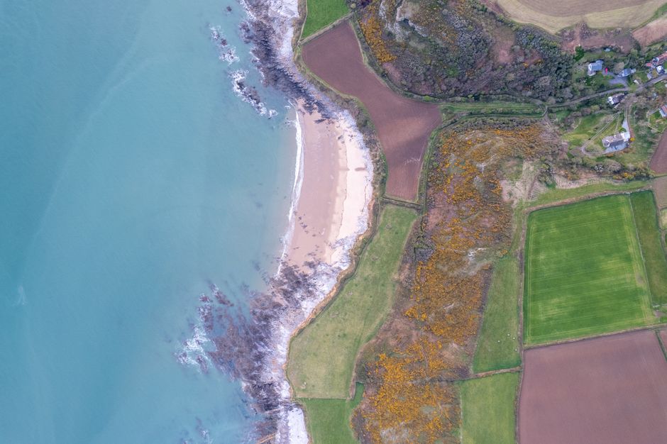 Aerial view of a coastal area showing a sandy beach with light-colored sand and dark rocks along the shoreline, where gentle waves are lapping against the coast. Behind the beach, there are patches of green fields and wooded areas with some trees displaying autumnal foliage. A narrow pathway runs through the fields, leading to a few small buildings located in the top right corner of the image. The scene is captured during daylight with natural lighting, highlighting the contrast between the ocean, land, and vegetation. This landscape is part of a rural or semi-rural setting, suitable for house relocations or moving services that involve transferring belongings through scenic environments. The image provides a clear view of the natural surroundings and the arrangement of the land, which may be relevant for planning furniture transport or home relocation logistics in the area served by Man with Van Slade Green.
