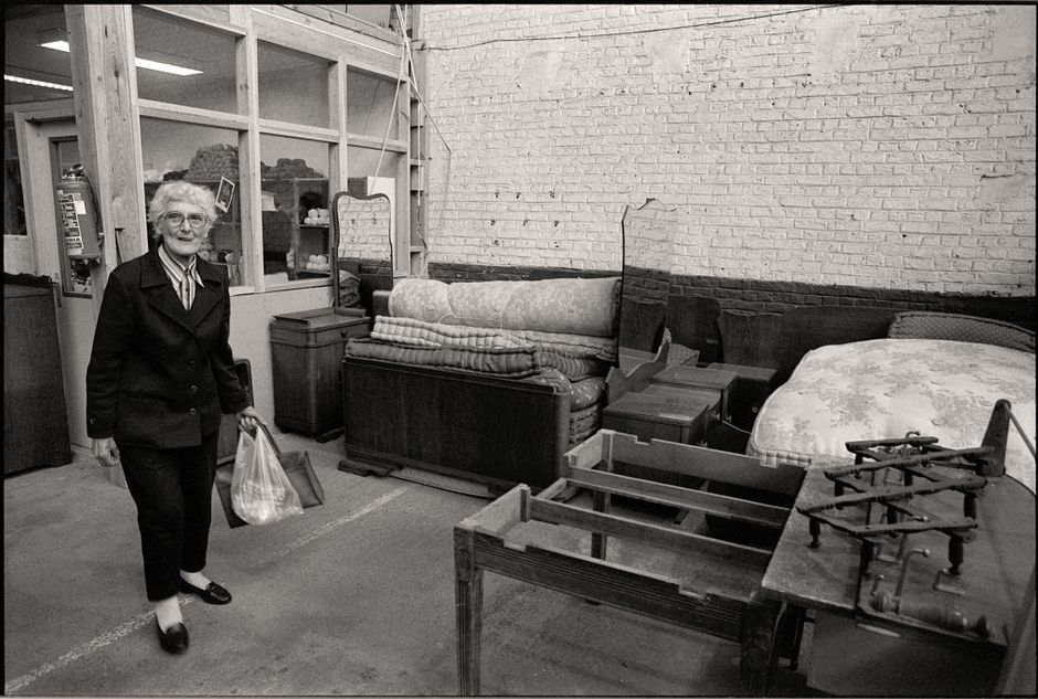 Image showing an elderly woman inside a storage or furniture removal setting, standing on a concrete floor and holding a plastic shopping bag in her right hand and a smaller handbag in her left hand. Behind her, there are various pieces of furniture including a stack of rolled-up carpets placed on a wooden sofa frame, a bed base with a mattress cover, and a small wooden bedside table. To the right, a two-burner portable gas stove is positioned on a wooden surface. The background features a white brick wall partially painted black, and a large window or partition with glass panels, indicating the space may be used for home relocation or furniture transport activities. The scene is lit with natural or overhead lighting, emphasizing the arrangement of furniture and packaging materials prepared for a moving or packing process. This image illustrates the type of objects and environment involved in house removals, as handled by Man with Van Slade Green.