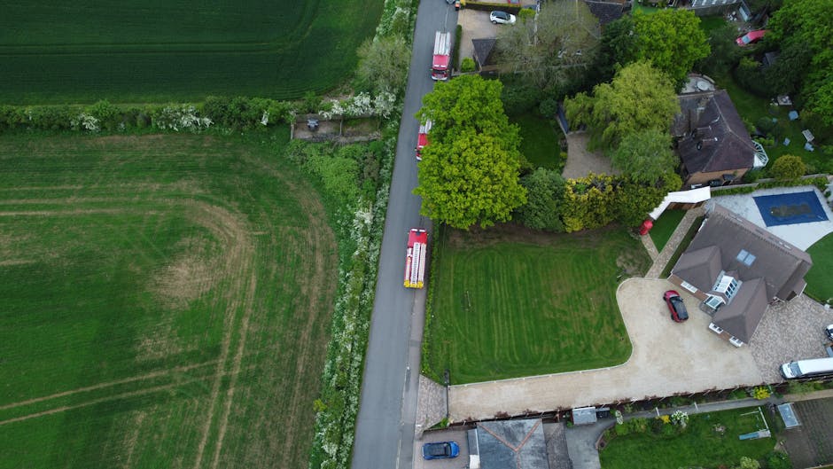 Aerial view of a residential property showing a narrow driveway with a red and white removals van parked at the entrance, adjacent to neatly mowed green lawns and surrounding mature trees. The propert