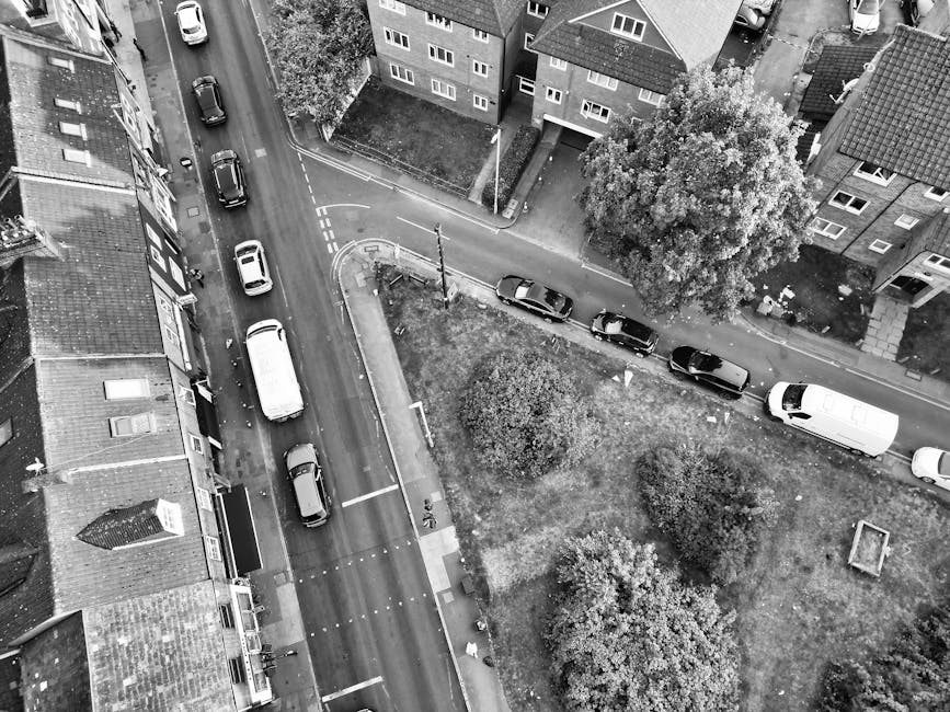 A black-and-white aerial photograph shows a narrow residential street with several parked cars along the curb, including a white van positioned near the edge of the street. The street is lined with multi-storey houses made of brick, with pitched roofs and multiple windows, some with front gardens or small yards. There are a few larger trees and bushes scattered along the pavement, providing some greenery to the scene. At the top of the image, a lane exit is visible, with a slight curve leading to the main road. Near the center, a small patch of grass and planting area is surrounded by a low curb, with a few pedestrians visible walking nearby. Inside a property, partially visible, are cardboard boxes and packing materials, possibly in the process of being packed or unloaded as part of a home relocation or furniture transport operation. The background features more houses and parked vehicles, with some suggesting ongoing loading or unloading activities, typical of a house removals or moving process. Man with Van Slade Green would use such scenes to illustrate their local household moving and packing services.