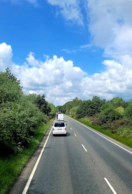A clear daytime scene of a two-lane suburban road with a row of green trees and shrubs on each side, leading into a distance under a partly cloudy sky with large white clouds and patches of blue. Several vehicles, including a small white van, are visible traveling along the road, which appears to be part of a home relocation process involving furniture and belongings being transported. The road surface is asphalt with white dashed lane markings, and the vehicles are spaced out evenly as they move forward. The natural surroundings and the open sky create an environment suitable for timely and organized moving logistics undertaken by professionals such as Man with Van Slade Green, who specialise in removals and furniture transport services.