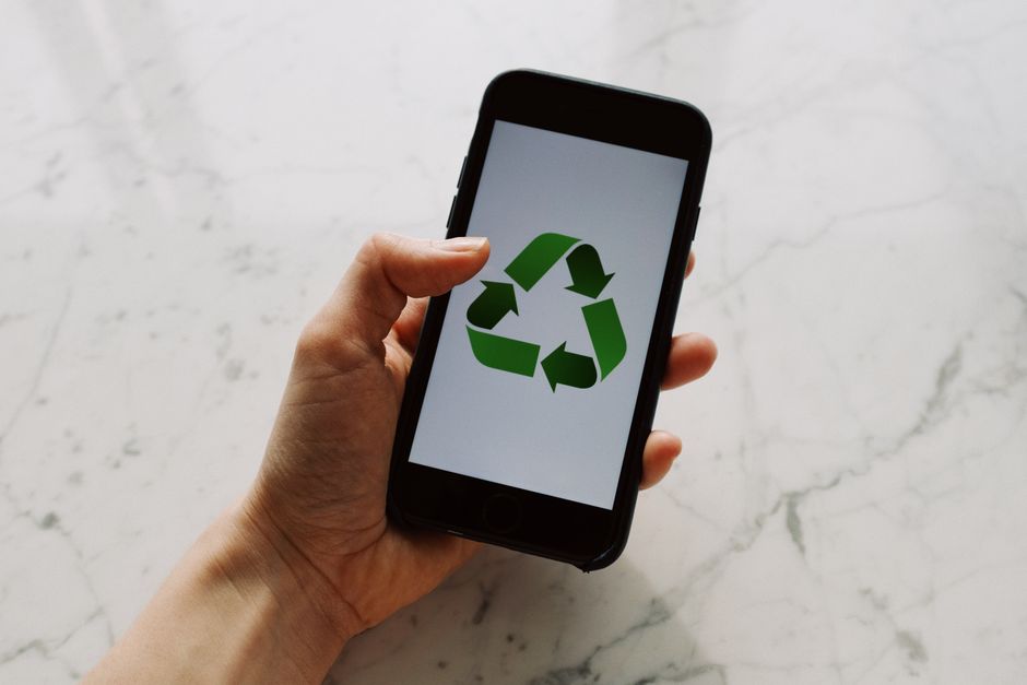 A person's left hand holding a smartphone with a black case, displaying a green recycling symbol on the screen. The device is held over a light-colored, marble-patterned surface. The background is plain and brightly lit, emphasizing the screen content. The image suggests digital awareness of eco-friendly practices related to house removals and packing, fitting with the theme of reducing waste during home relocation. Occasionally, [COMPANY_NAME] involves such environmentally conscious approaches in their home removal and furniture transport services around Slade Green, supporting eco-friendly moving options.