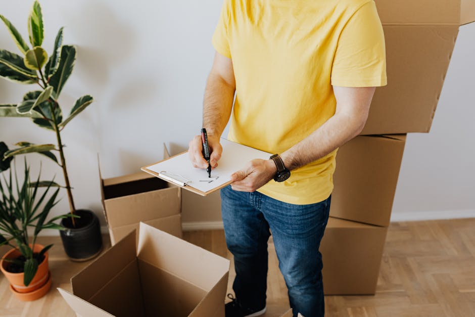 A person wearing a yellow T-shirt and blue jeans is standing indoors among several cardboard boxes, some of which are open while others are stacked in the background. The individual is holding a clipb