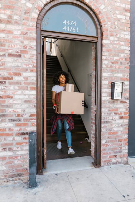 A young woman with curly hair carrying a medium-sized cardboard box and a larger box at her waist level, descending the stairs through the open entrance of a brick building. She is dressed in casual clothing, including a white t-shirt, blue jeans, white sneakers, and has a red plaid shirt tied around her waist. Behind her, the interior staircase, made of dark material, ascends inside the building. The entrance features an arched window pane above the door with the numbers 4474 and 4476 displayed, and a doorbell or intercom device is mounted on the brick wall to the right of the door frame. This scene is part of a home relocation or moving process, possibly coordinated by Man with Van Slade Green, emphasizing careful packing and furniture transport during an internal or external load transfer for house removals at Slade Green Station flats.