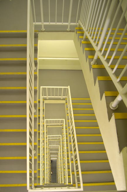 A view of a multi-story concrete stairwell inside a residential building, showing a series of descending flights with white metal railings on both sides. The stairs have yellow safety strips along the edge of each step, providing traction and visibility. The staircase appears well-lit with artificial lighting, and the downwards perspective emphasizes the depth and repetitive pattern of the stairs. The environment is clean and orderly, suitable for the transport of household items during a home relocation or moving process. This image supports content related to moving tips for flats, such as those at Slade Green Station, and illustrates the internal staircase used by movers like Man with Van Slade Green during furniture transport or packing and moving tasks.