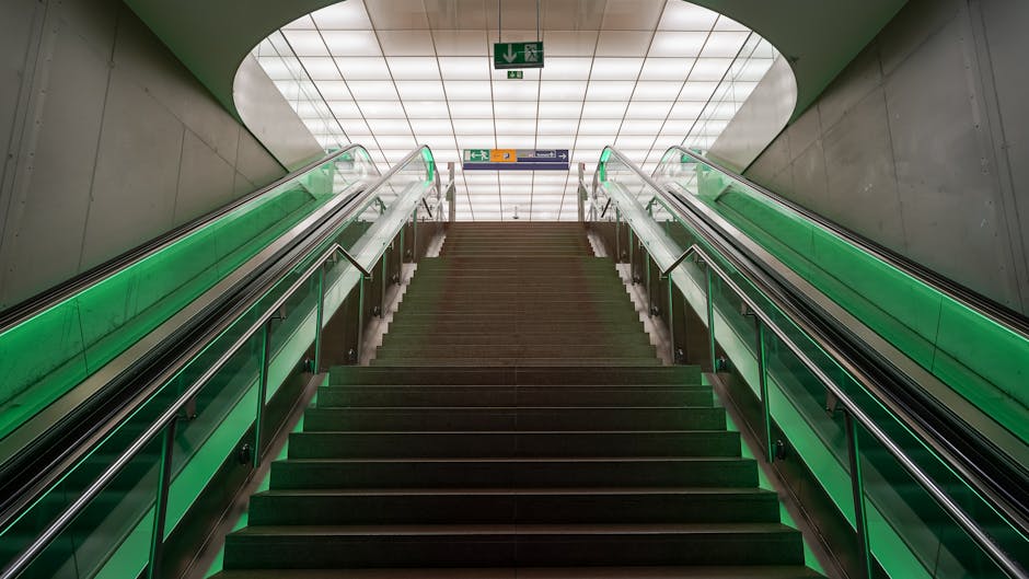A young woman with curly hair carrying a medium-sized cardboard box and a larger box at her waist level, descending the stairs through the open entrance of a brick building. She is dressed in casual clothing, including a white t-shirt, blue jeans, white sneakers, and has a red plaid shirt tied around her waist. Behind her, the interior staircase, made of dark material, ascends inside the building. The entrance features an arched window pane above the door with the numbers 4474 and 4476 displayed, and a doorbell or intercom device is mounted on the brick wall to the right of the door frame. This scene is part of a home relocation or moving process, possibly coordinated by Man with Van Slade Green, emphasizing careful packing and furniture transport during an internal or external load transfer for house removals at Slade Green Station flats.